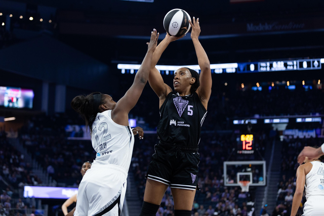 A basketball player in a black jersey is shooting the ball while a player in a white jersey attempts to block. The shot clock shows 12 seconds left.